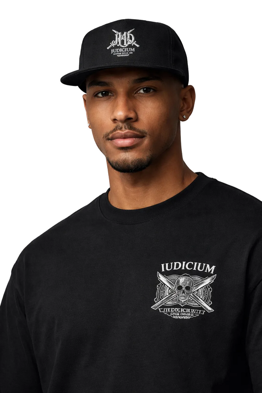 Man wearing a black cap and black sweatshirt with 'IUDICUM' logo on a gray background