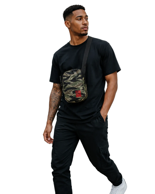 A man wearing a black t-shirt, black pants, and white sneakers walks against a concrete wall while wearing a camouflage crossbody bag with a red logo patch.