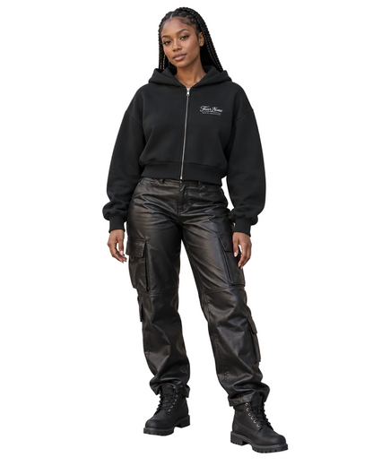 A woman stands on a city sidewalk wearing a black cropped zip-up hoodie with a small white chest logo, black leather cargo pants, and black lace-up boots, posed against a beige stone building.