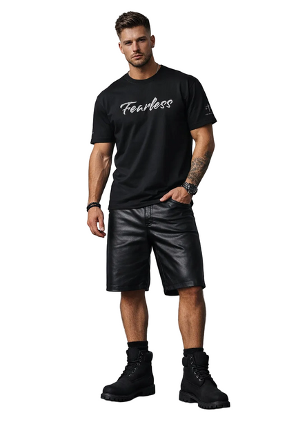 Man wearing a black t-shirt with 'Fearless' text, black leather shorts, and black boots against a gray brick wall.