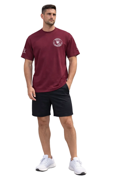 Man wearing a maroon t-shirt with a logo and black shorts, standing in front of a gray building.