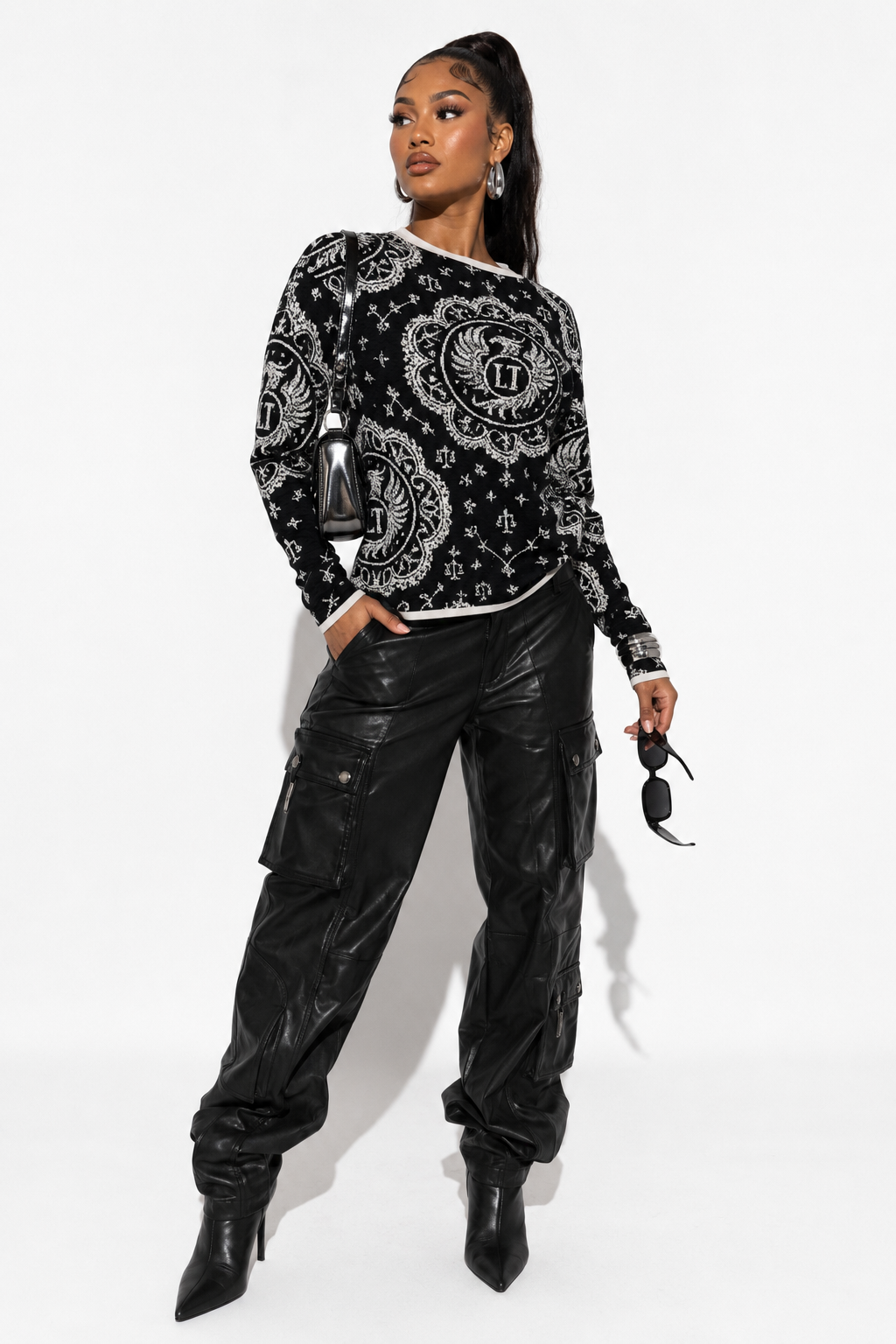 A model wearing a black and white paisley patterned long-sleeve top, black faux-leather cargo pants, and black pointed-toe heeled boots, holding sunglasses against a white studio background.
