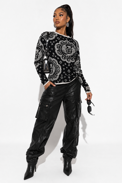 A model wearing a black and white paisley patterned long-sleeve top, black faux-leather cargo pants, and black pointed-toe heeled boots, holding sunglasses against a white studio background.