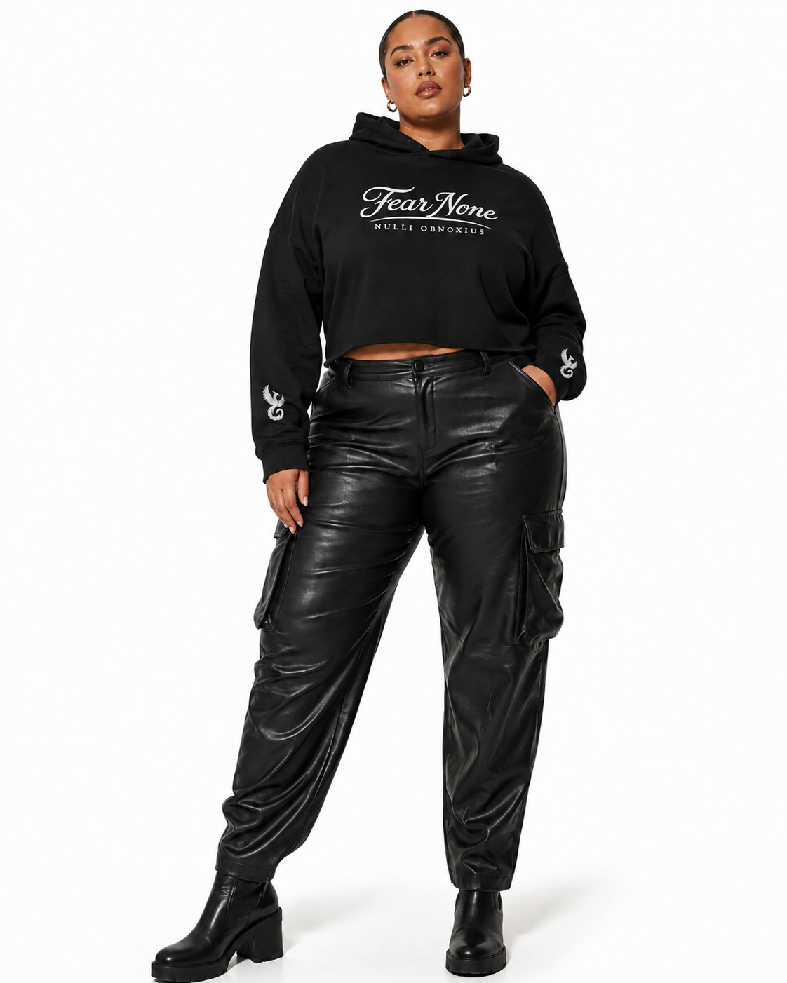 A model wearing a black Fear of God New York crop hoodie with white logo text and black faux-leather cargo pants, standing against a plain white background.