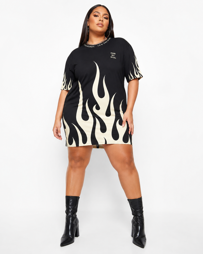 A plus-size model wearing a black short-sleeve mini dress with a bold cream flame pattern and black pointed-toe ankle boots, standing against a plain white studio background.