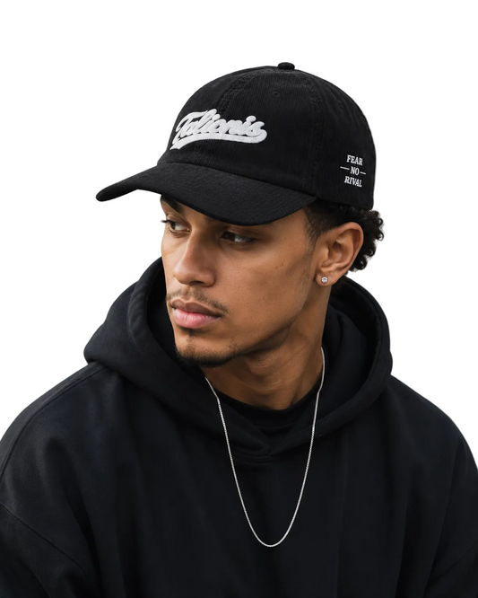 A man wearing a black baseball cap with white embroidered 'Clerks' branding on the front, paired with a black hoodie and a silver chain necklace, set against a concrete wall background.