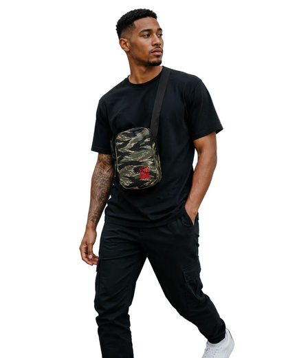 A man wearing a black t-shirt, black pants, and white sneakers walks against a concrete wall while wearing a camouflage crossbody bag with a red logo patch.