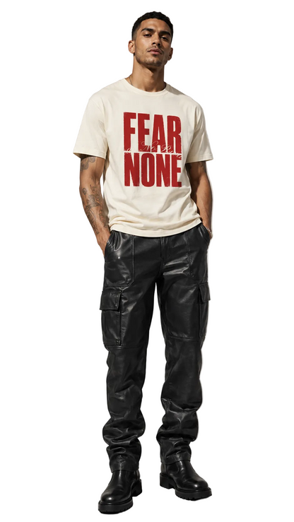 A man modeling a cream-colored t-shirt with bold red text reading 'FEAR NONE' on the front, paired with black cargo pants and black boots, against a neutral studio background.