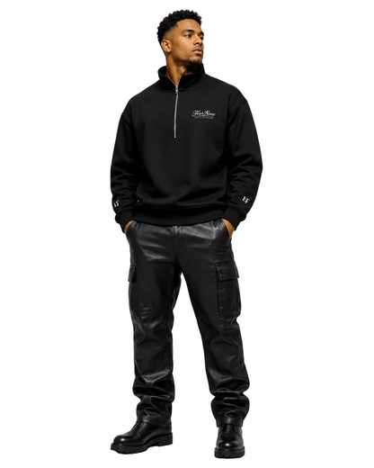 A man wearing a black half-zip sweatshirt with a small white chest logo, black cargo pants, and black boots, standing against a light gray studio background.