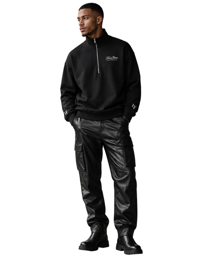 A man wearing a black half-zip pullover sweatshirt with a small white chest logo, black cargo pants, and black boots, standing in a modern indoor setting with gray and black marble walls.