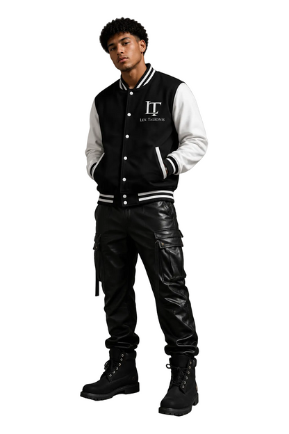 Person wearing a black and white varsity jacket with a logo, black pants, and black boots against a gray background.