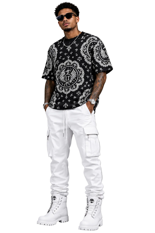 Man wearing a black patterned shirt, white pants, and white boots against a gray background