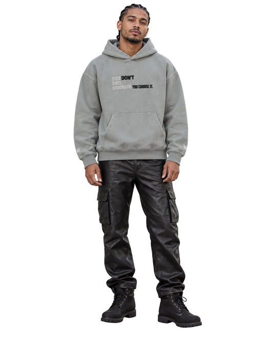A man stands outdoors on a sidewalk wearing a light gray pullover hoodie with black text on the chest, black leather cargo pants, and black boots, posed in front of a light-colored concrete building.
