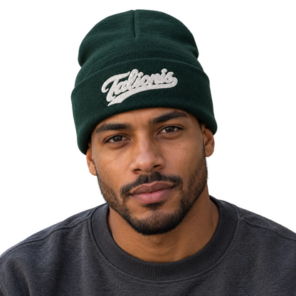 A man wearing a dark green knit beanie with 'Talionis' embroidered in white script, photographed outdoors against a light stone wall.